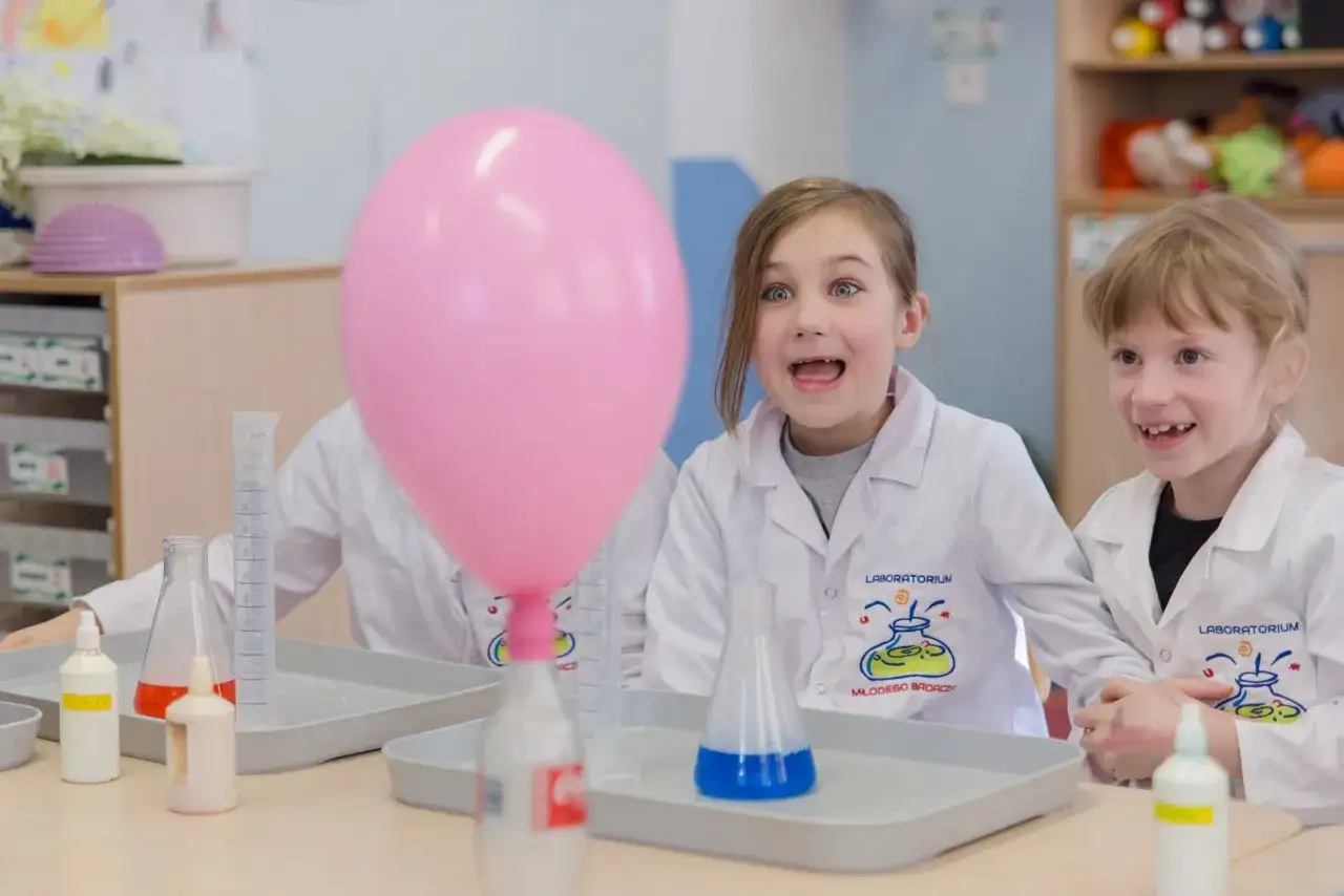 Dzieci w fartuchach laboratoryjnych z ekscytacją obserwują różowy balon podczas eksperymentów chemicznych dla dzieci.