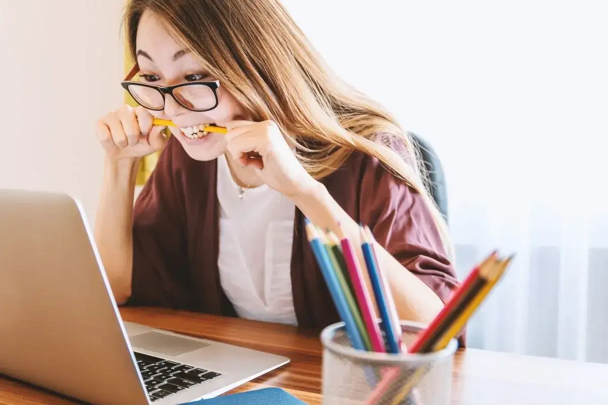 Kobieta w okularach gryzie ołówek, pracując przy laptopie. Kolorowe kredki w pojemniku sugerują kreatywność, ale jej mina zdradza trudności w osiągnięciu work life balance.