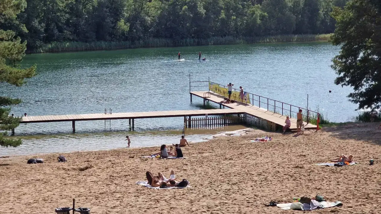 Słoneczna borówno plaża nad jeziorem, ludzie odpoczywają na piasku i w wodzie, niektórzy pływają na desce SUP.