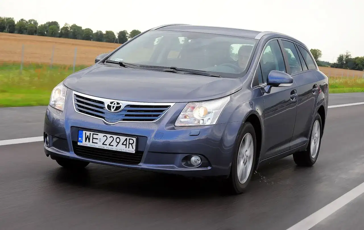 Szary kombi Toyota Avensis T27 1.8 benzyna jedzie po drodze, za nim pola i drzewa.