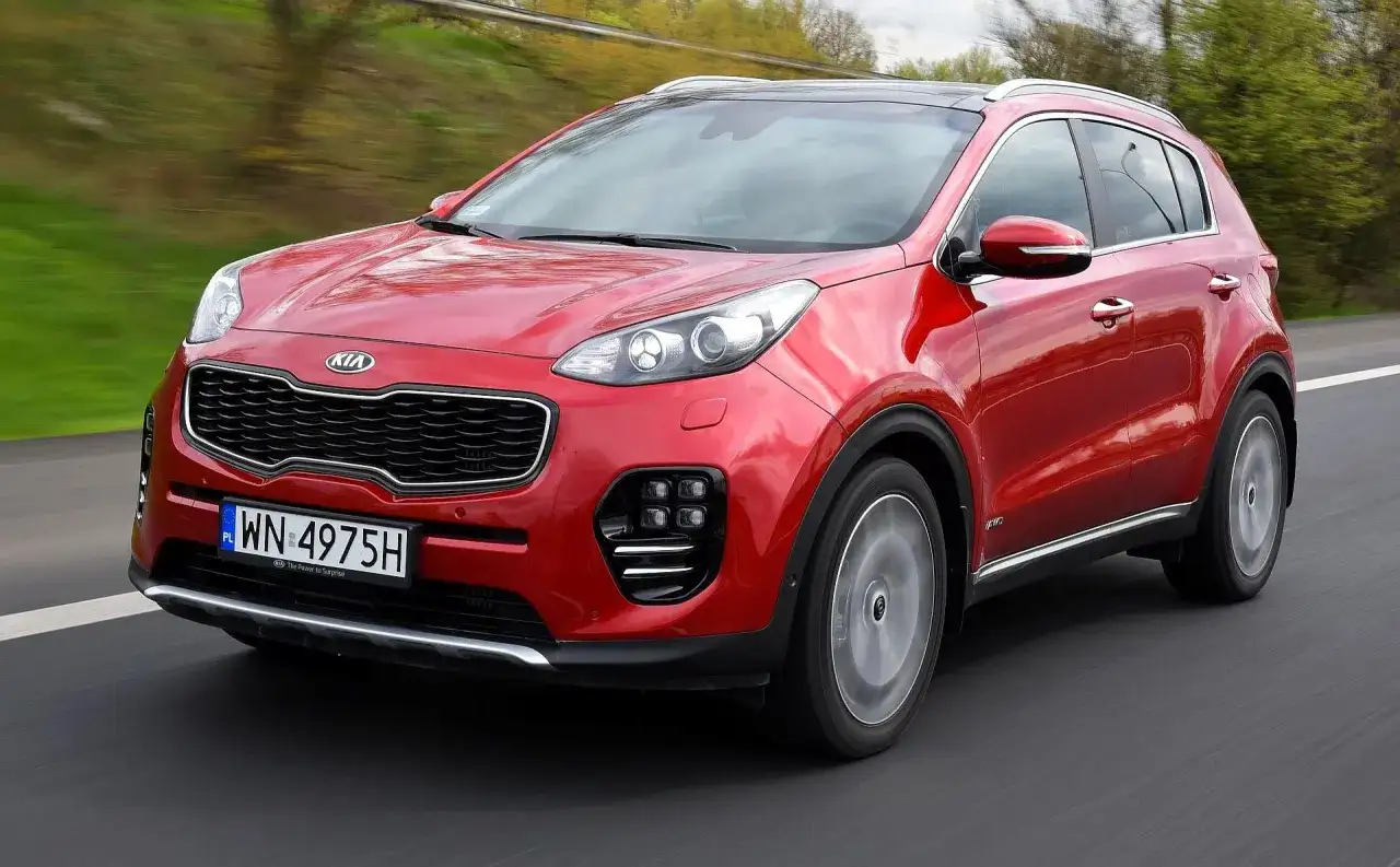 Czerwony Kia Sportage IV pędzi po drodze, otoczony zielenią.