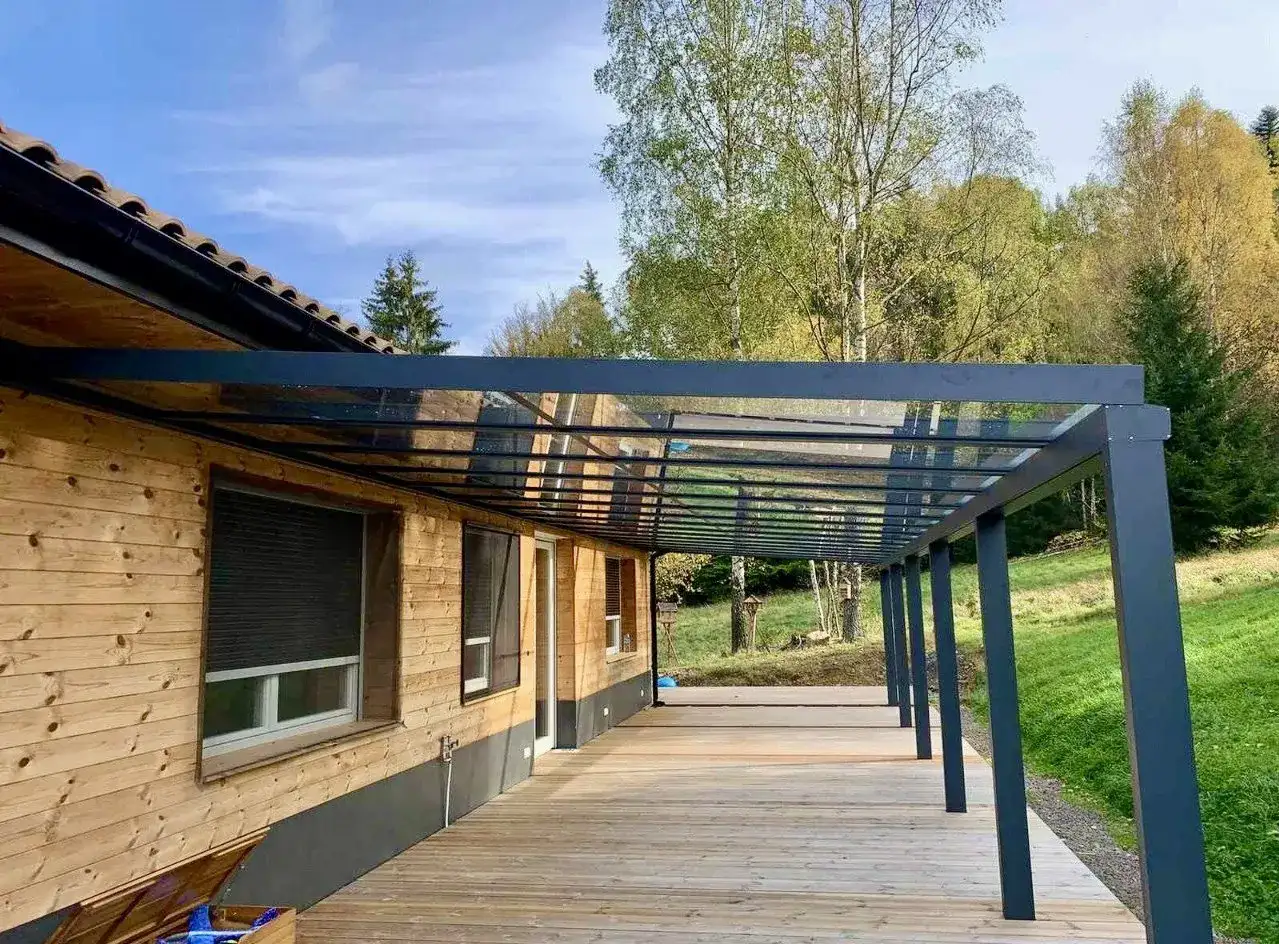Drewniany taras pod dachem z przezroczystego tworzywa, przylegający do domu z drewnianą elewacją. Otoczony zielenią i drzewami.