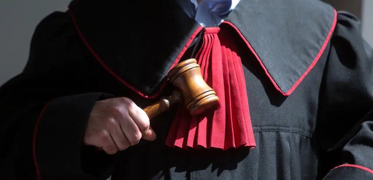 Sędzia trzyma młotek, gotowy do ogłoszenia wyroku. W tle widać togę z czerwonym żabotem, symbolizującą sprawiedliwość i powagę, która towarzyszy wydaniu subsydiarnego aktu oskarżenia.