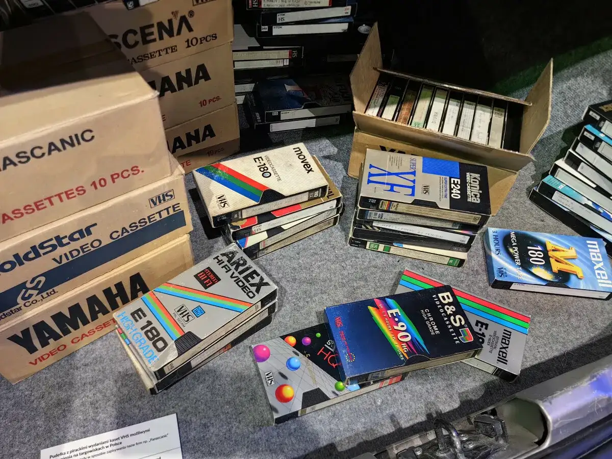 Kolekcja kaset VHS, w tym marki Movix, Konica, Maxell, Ariex, Yamaha. W tle pudełka z kasetami, przypominające o opłacie reprograficznej.
