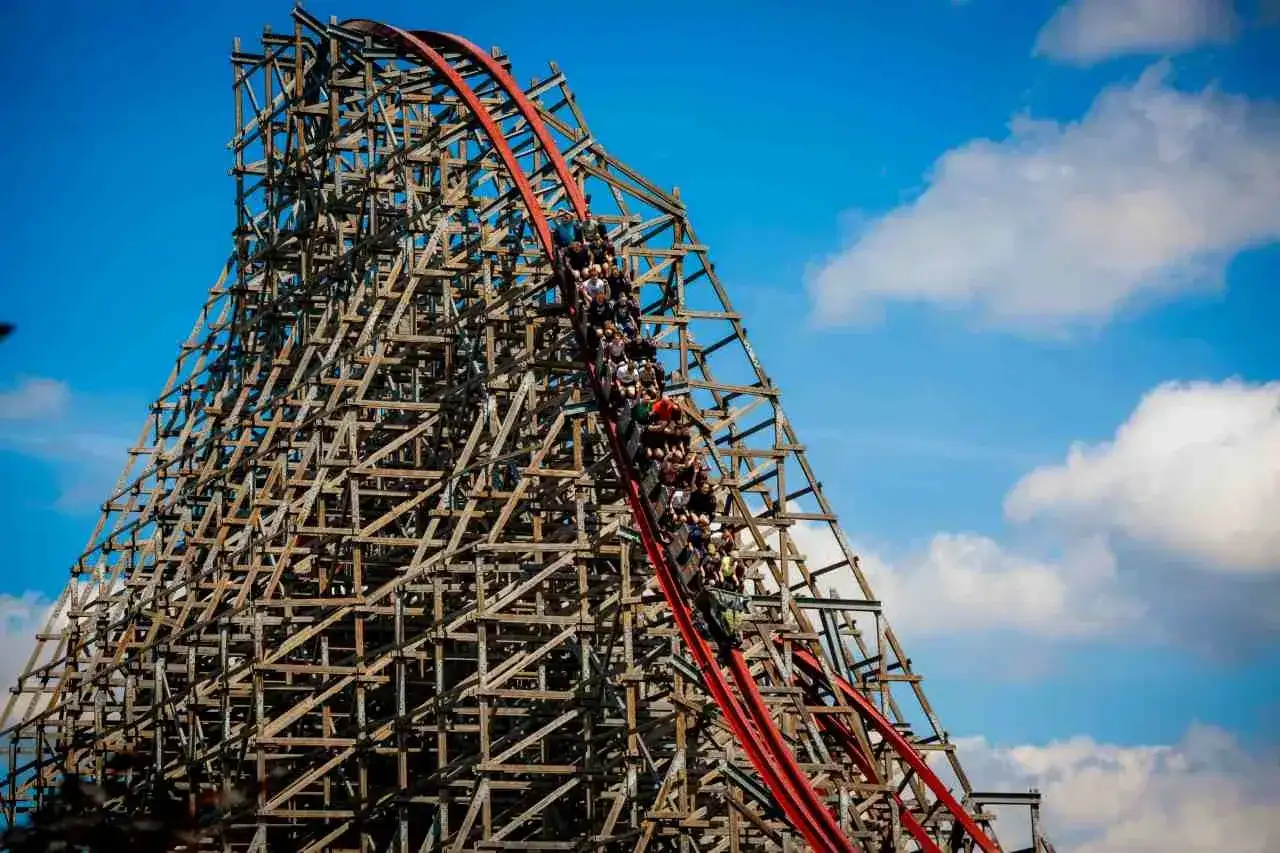 Pasażerowie Hyperion roller coaster zjeżdżają po drewnianej konstrukcji, która wznosi się ku błękitnemu niebu z chmurami.
