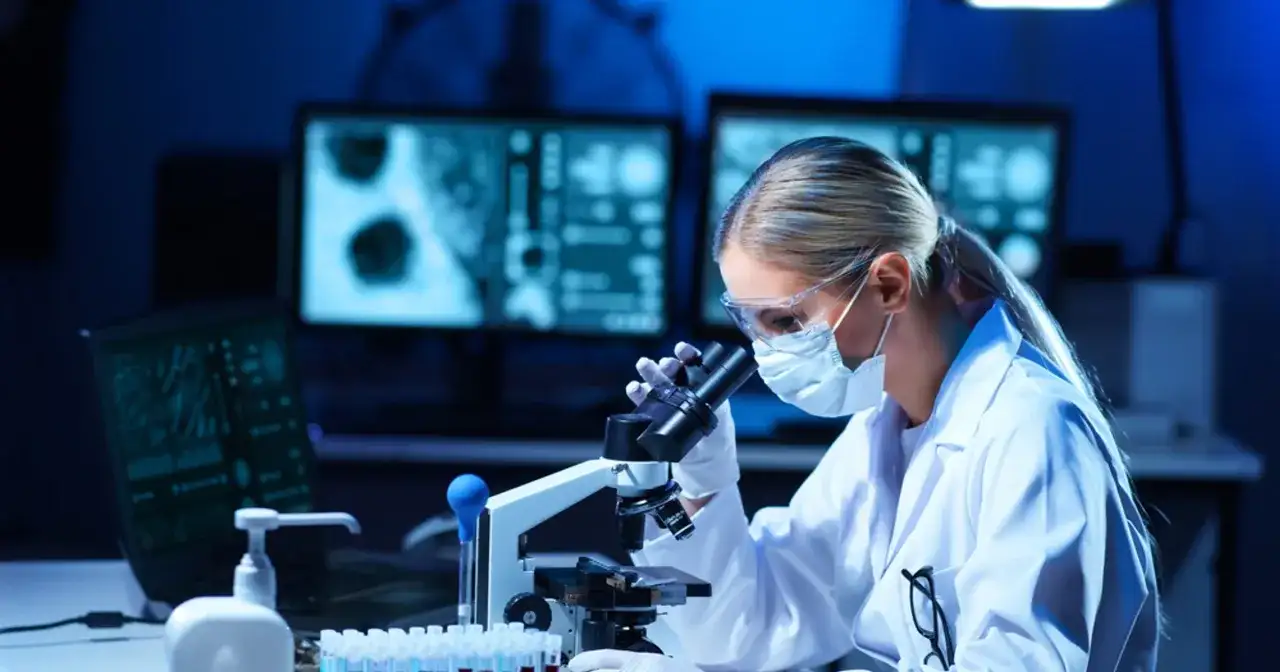 Diagnosta laboratoryjny w okularach i masce pochyla się nad mikroskopem, analizując próbki.