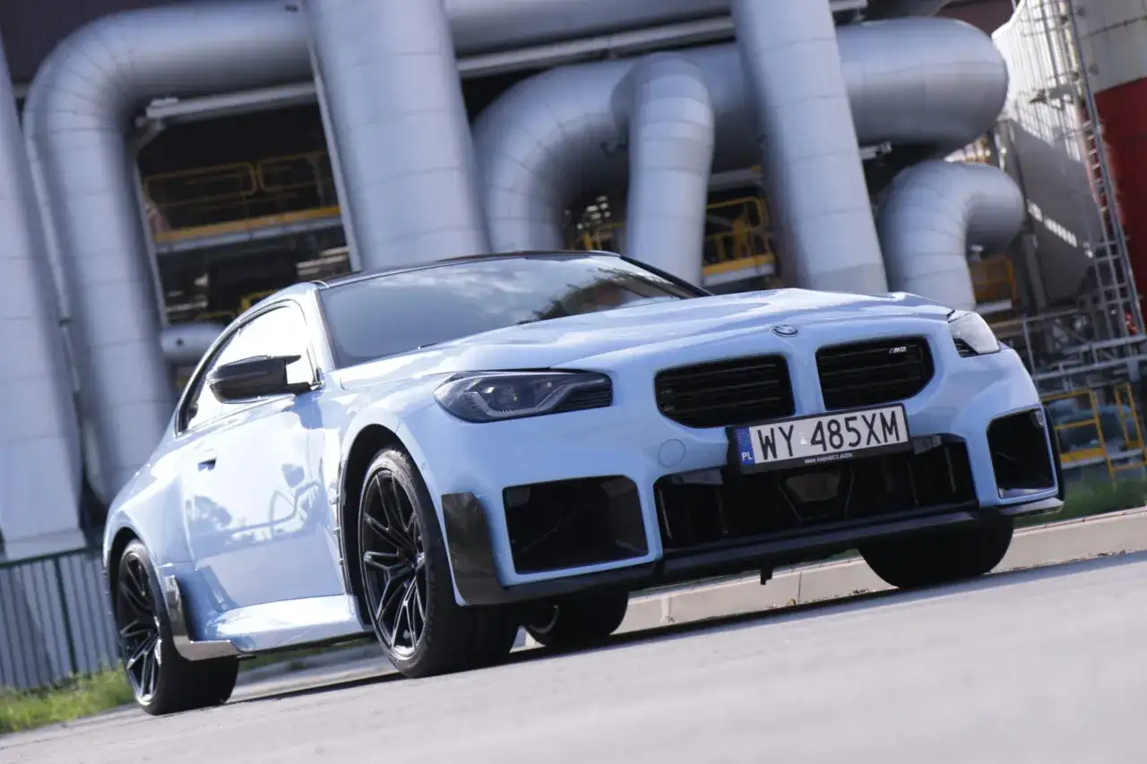 Jasnoniebieskie BMW M2 z czarnymi felgami i spoilerami, na tle przemysłowych rur. Sprawdź aktualną bmw m2 cena.