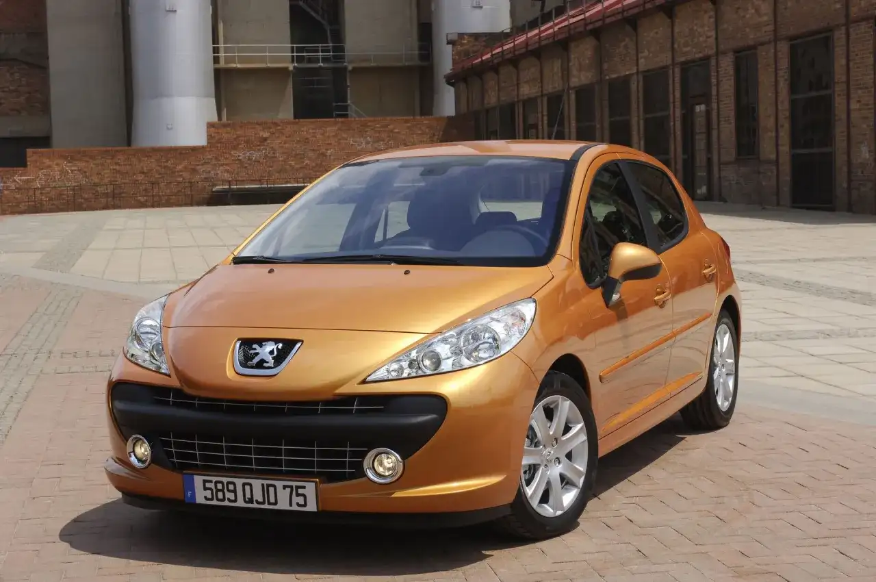 Pomarańczowy Peugeot 207 SW 1.4 benzyna zaparkowany na brukowanym placu przed ceglanym budynkiem.