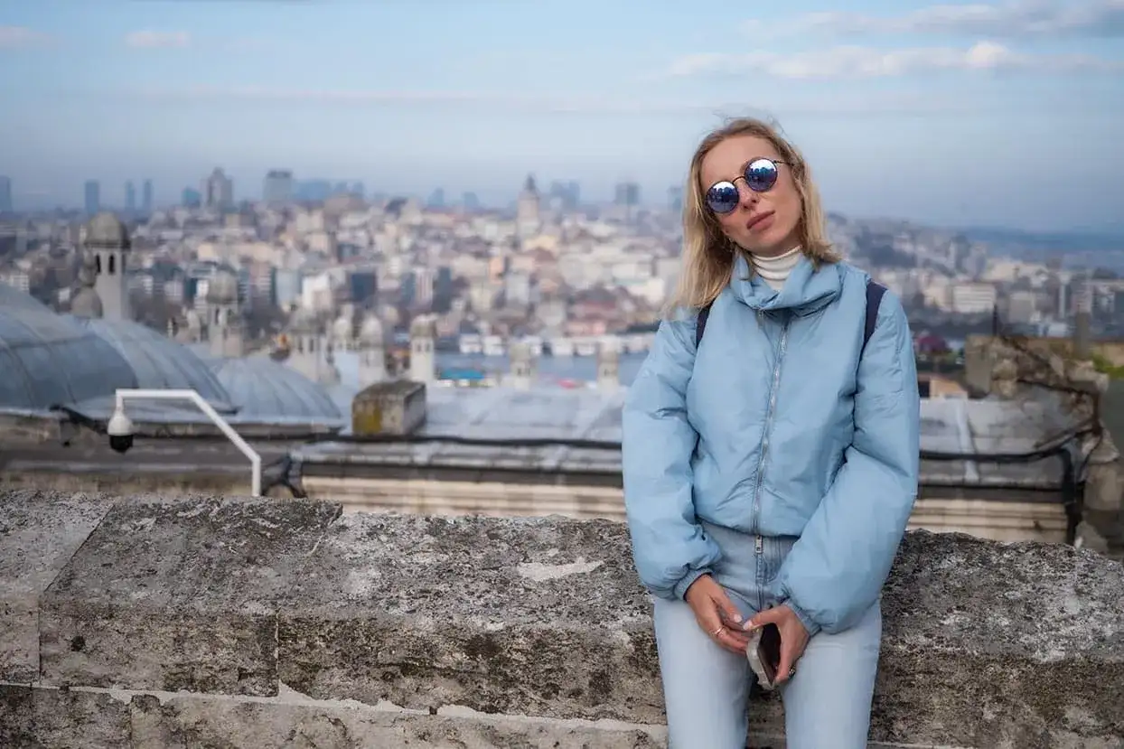 Kobieta w jasnoniebieskiej kurtce i okularach przeciwsłonecznych na tle panoramy Stambułu.