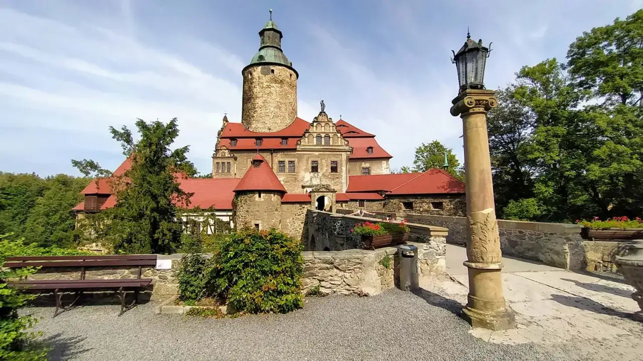 Zamek Czocha, gdzie jest? Malowniczy zamek z czerwoną dachówką i wieżą, otoczony zielenią, z kamiennym mostkiem i latarnią.