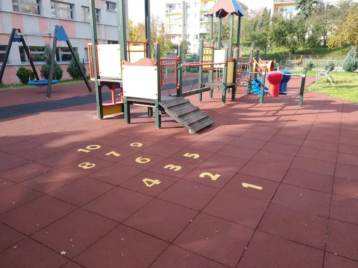 Plac zabaw z huśtawkami i drabinkami. Na czerwonej nawierzchni namalowana jest gra w klasy, a obok niej widać elementy do zabawy w twister.
