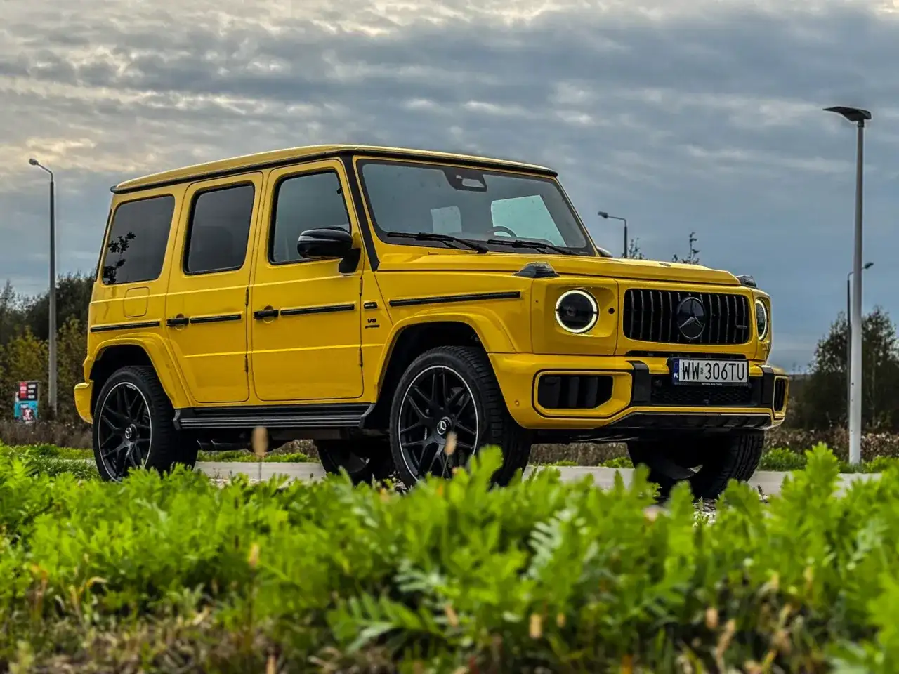 Żółty Mercedes G63 AMG z czarnymi felgami i detalami, zaparkowany na tle pochmurnego nieba i zieleni.