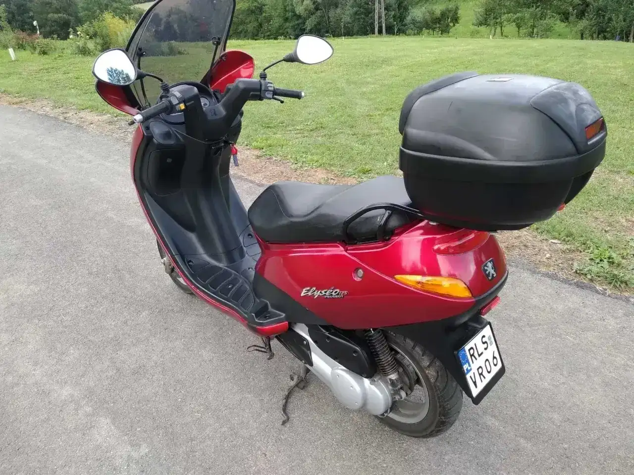 Czerwony skuter Peugeot Elyseo 125 z czarnym kufrem na bagaż, gotowy na podróż po malowniczych drogach.