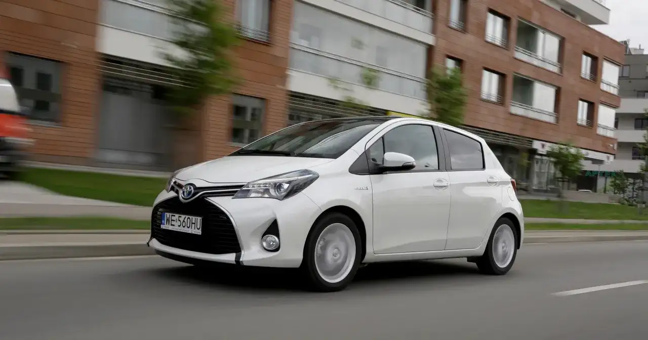 Biały model toyoty na 5 liter, Yaris Hybrid, jedzie ulicą obok nowoczesnych budynków.