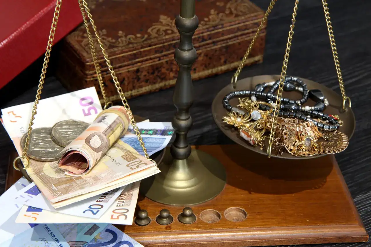 Waga szalkowa z euro i biżuterią. Na jednej szalce banknoty euro i monety, na drugiej złota biżuteria. Cena skupu 20 dolarów w złocie.