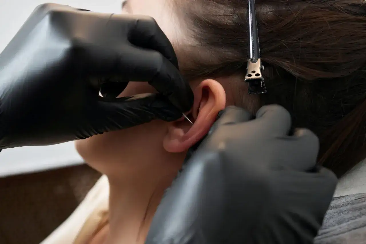 Profesjonalne wykonanie kolczyka orbital piercing. Ręce w czarnych rękawiczkach precyzyjnie przekłuwają ucho.