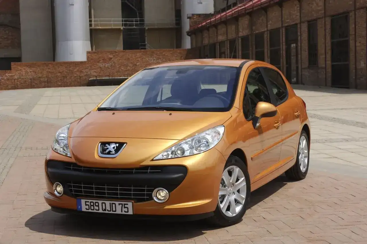 Pomarańczowy Peugeot 207 1.4 benzyna zaparkowany na placu z ceglaną posadzką, w tle budynki przemysłowe.