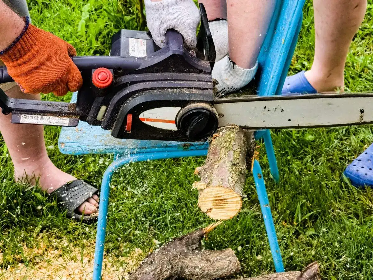 Osoba w pomarańczowej rękawicy używa piły łańcuchowej do cięcia drewna na prowizorycznym stojaku. To prosty sposób, jak zrobić stojak do cięcia drewna.