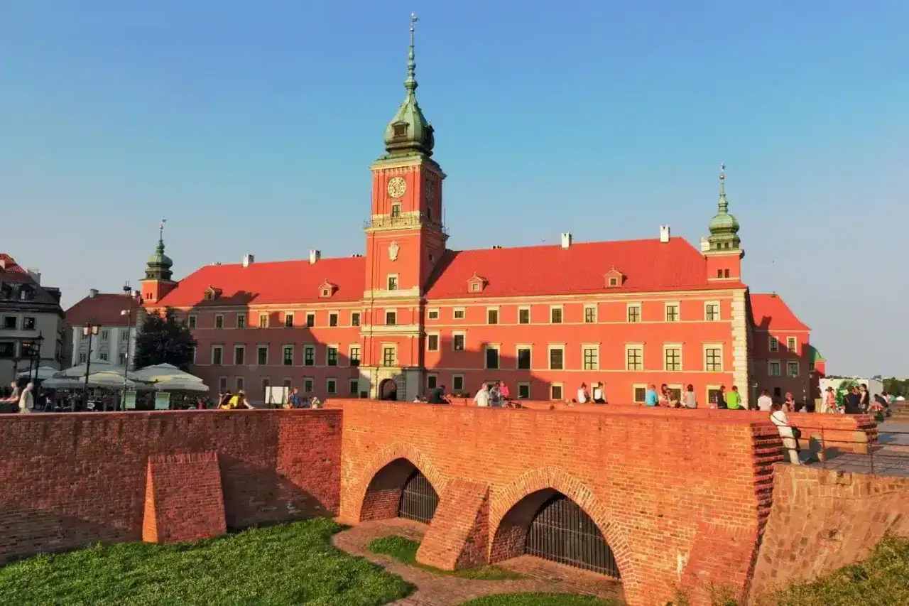 Zamek Królewski w Warszawie, z czerwoną fasadą i zieloną wieżą, widziany zza ceglanego mostu.