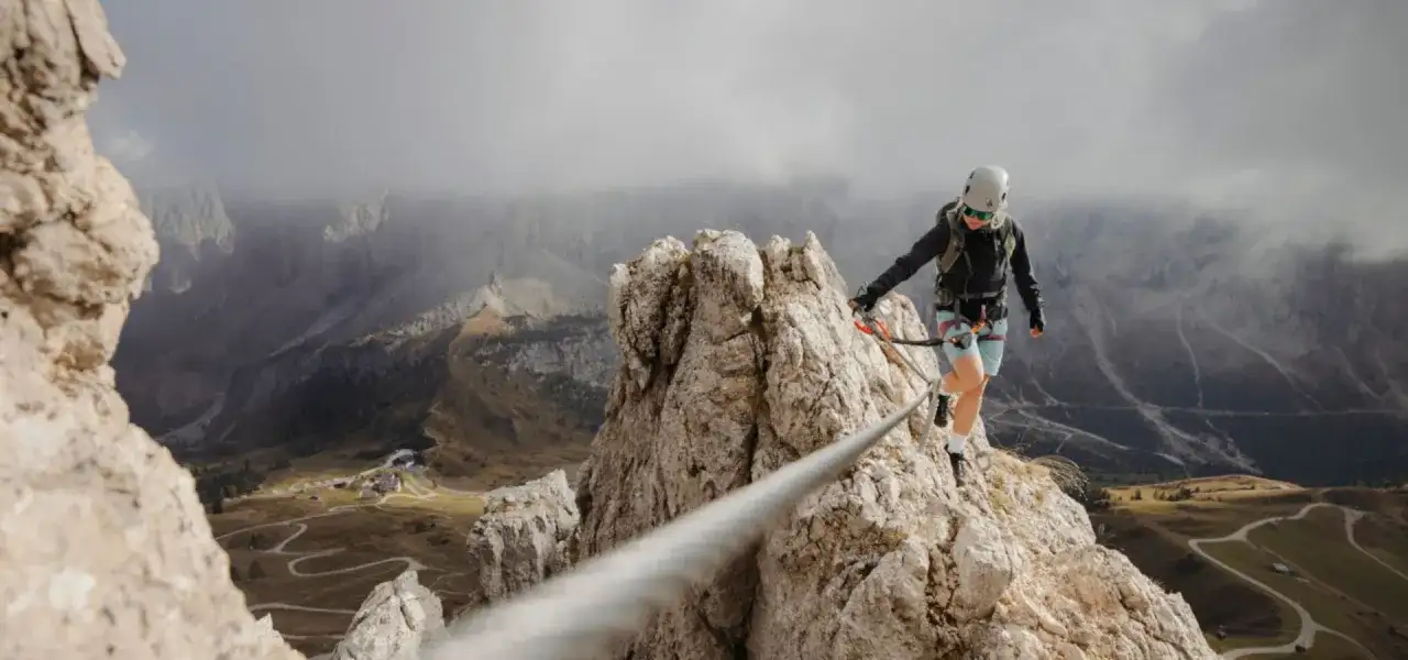 Kobieta na via ferrata polska pokonuje skalną grań, w tle górzysty krajobraz z krętymi drogami.