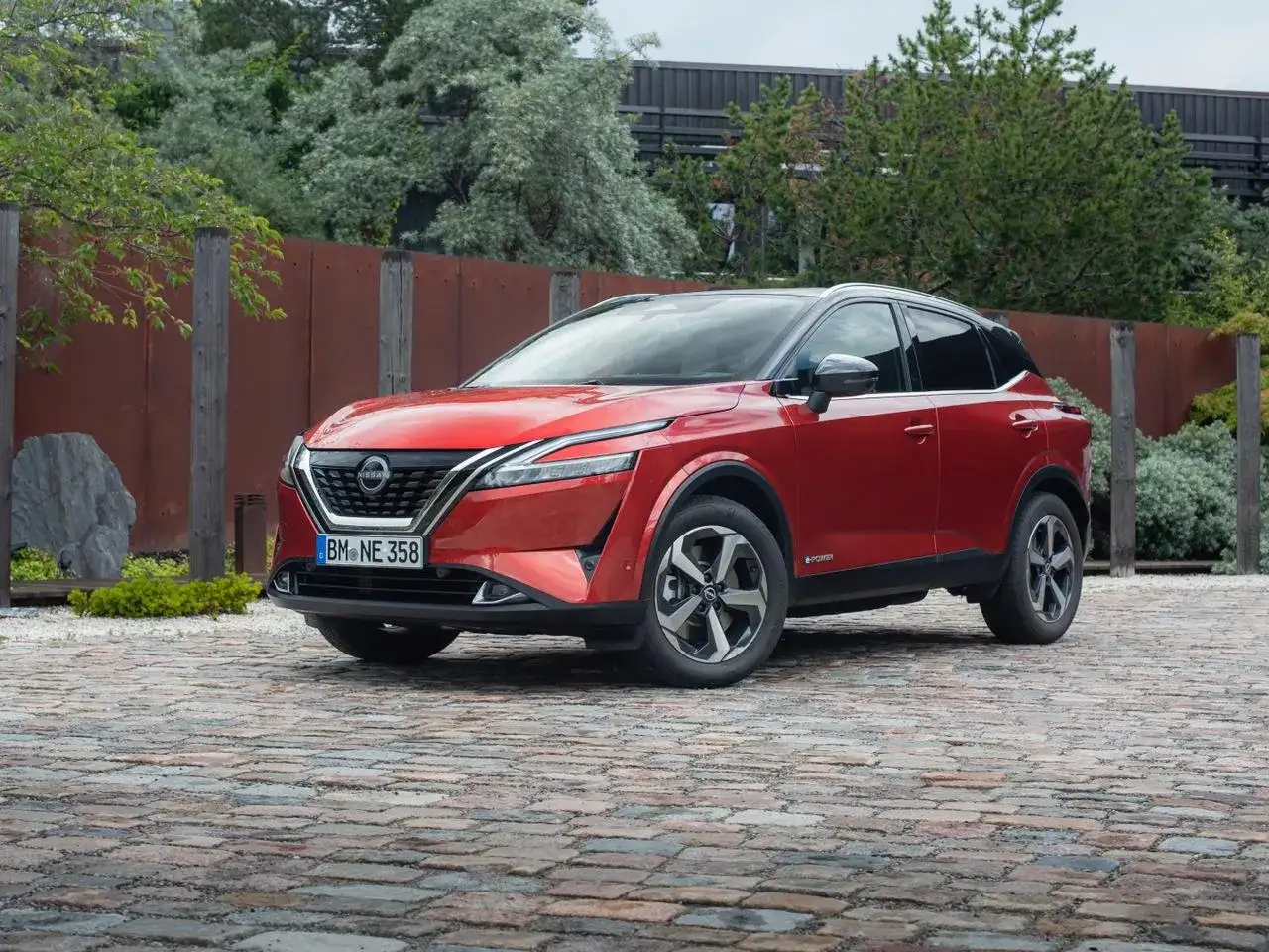 Czerwony Nissan Qashqai 2025 z czarnym dachem, zaparkowany na brukowanej nawierzchni, otoczony zielenią i industrialnym ogrodzeniem.