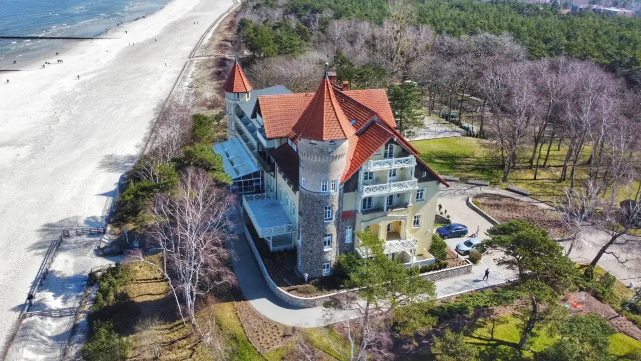Elegancki hotel z wieżą i czerwonym dachem oferuje noclegi Łeba blisko morza. Piękna plaża i las otaczają obiekt.