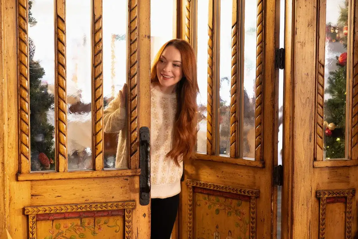 Lindsay Lohan w świątecznym swetrze zagląda przez ozdobne drzwi. W tle choinka, jak w jej filmach.