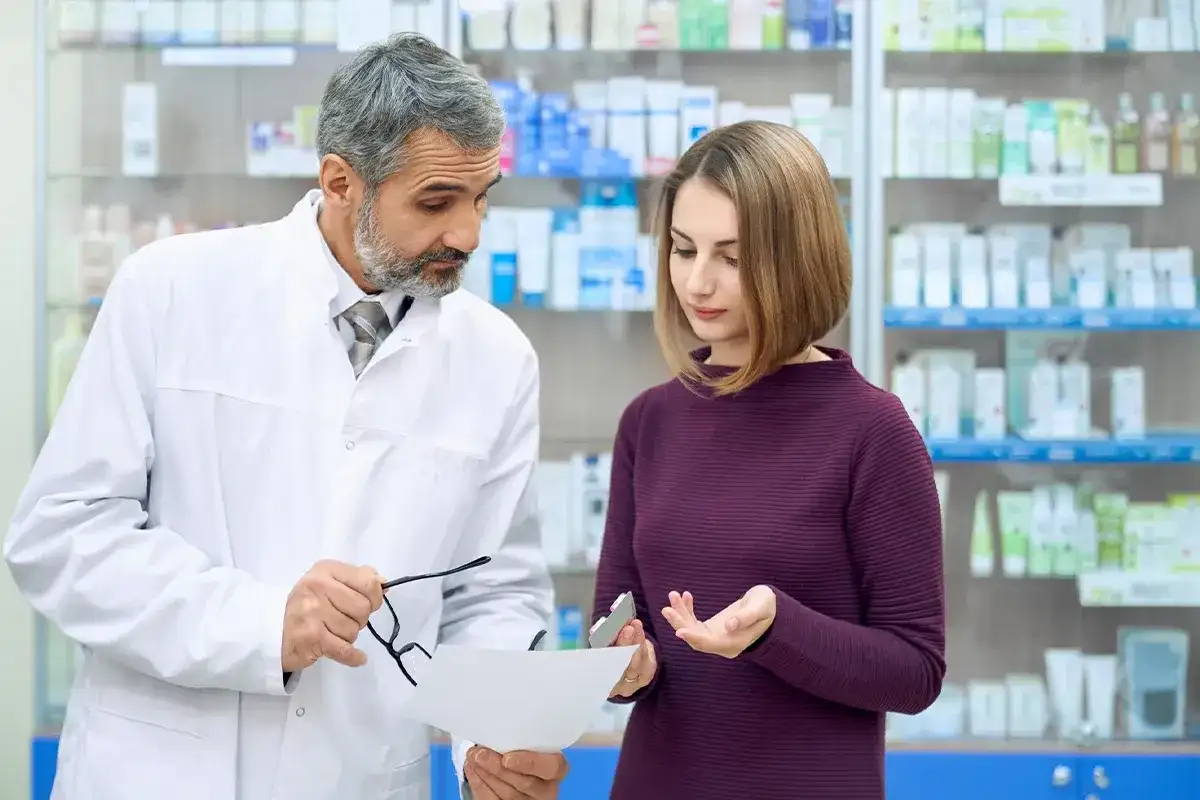 Doświadczony farmaceuta doradza klientce w aptece, wskazując na receptę i opakowanie leku.