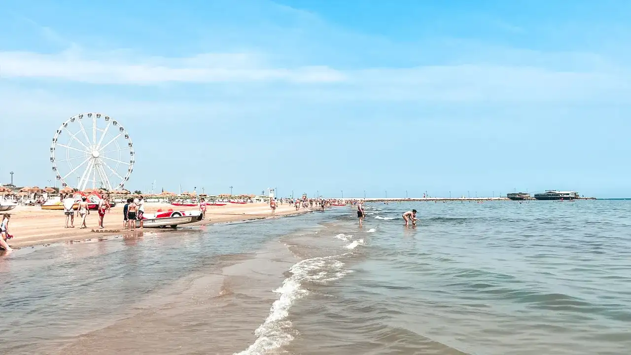 Słoneczny dzień na plaży w Rimini. Ludzie spacerują, bawią się w wodzie, a w oddali widać diabelski młyn. Idealne miejsce na wakacje!