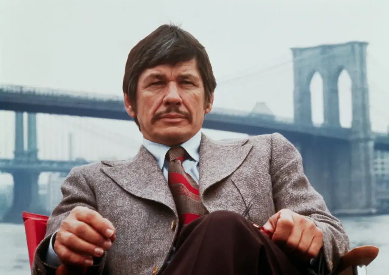 Charles Bronson w garniturze, z wąsami, siedzi przed mostem Brooklińskim. Ikona kina akcji, znany z wielu filmów.