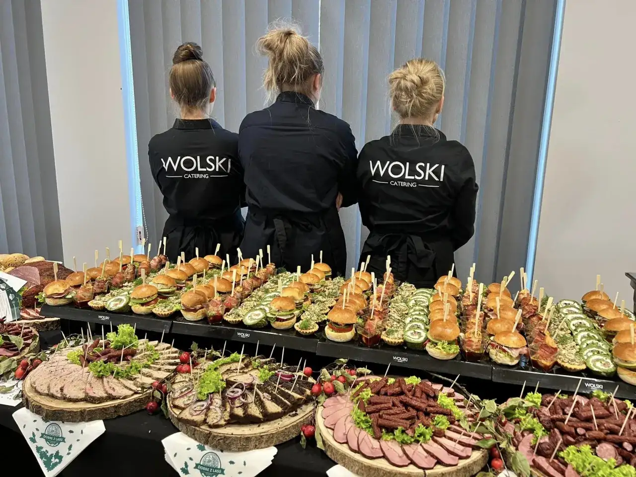 Trzy kobiety w czarnych fartuchach z logo "WOLSKI CATERING" stoją za stołem pełnym przekąsek. To idealna impreza z pysznym jedzeniem.