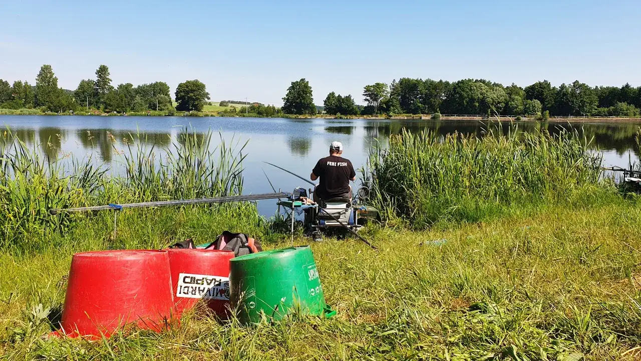 Wędkarz z napisem "FEBI FISH" na koszulce siedzi nad spokojnym stawem, gotowy do łowienia.