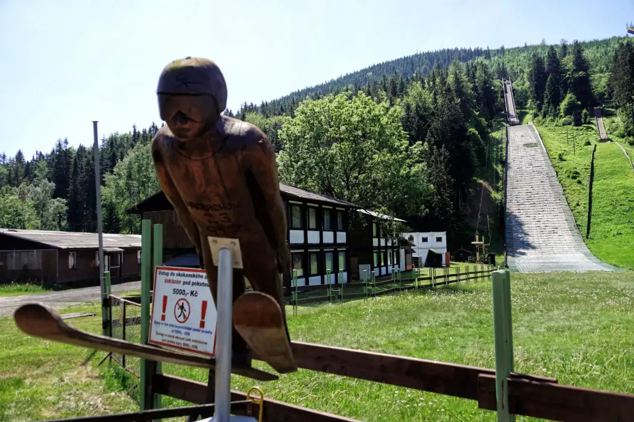 Drewniana figura skoczka narciarskiego przed skocznią w Harrachovie. W tle zielone wzgórza i lasy.