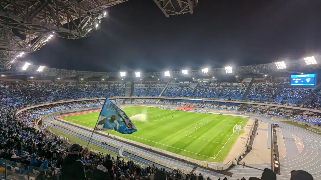 Widok na pełen kibiców stadion w Neapolu nocą. Trybuny wypełnione niebieskimi barwami, na murawie zielona trawa.