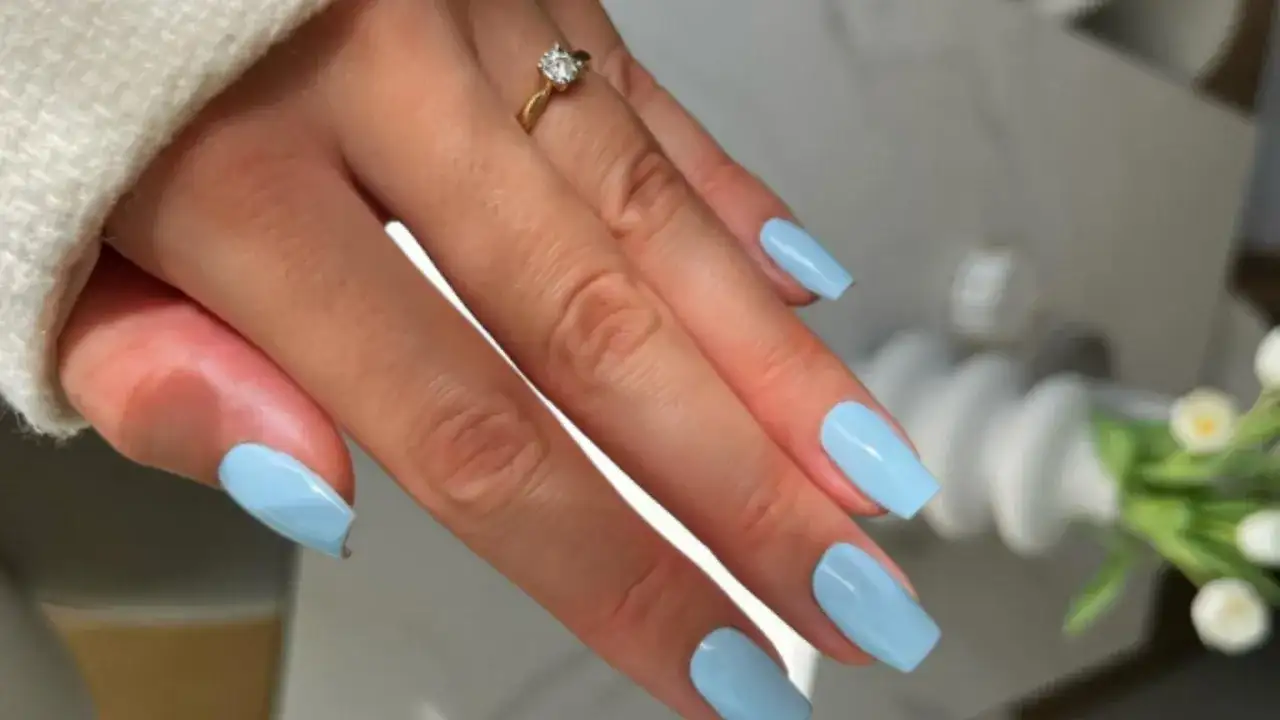Dłoń z paznokciami w kolorze baby blue, ozdobiona złotym pierścionkiem z brylantem.