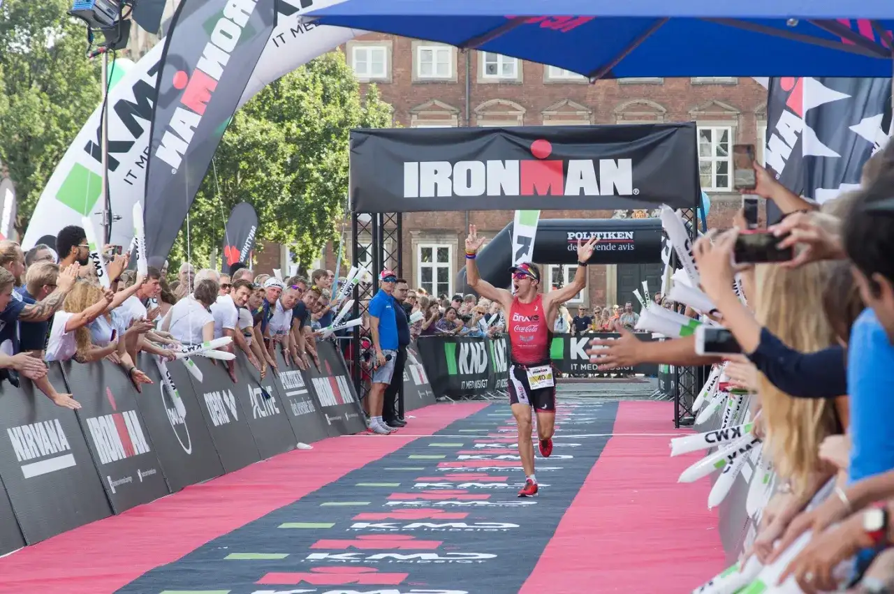 A triumphant athlete crosses the finish line at an Ironman triathlon, arms raised in victory, cheered by a crowd.