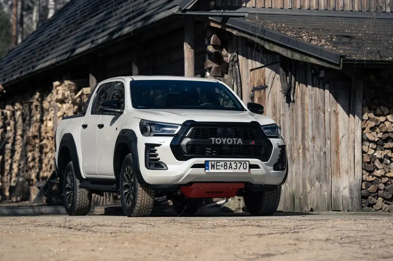 Biały Toyota Hilux GR Sport z czarnymi dodatkami, zaparkowany przed drewnianym budynkiem z polanami drewna.