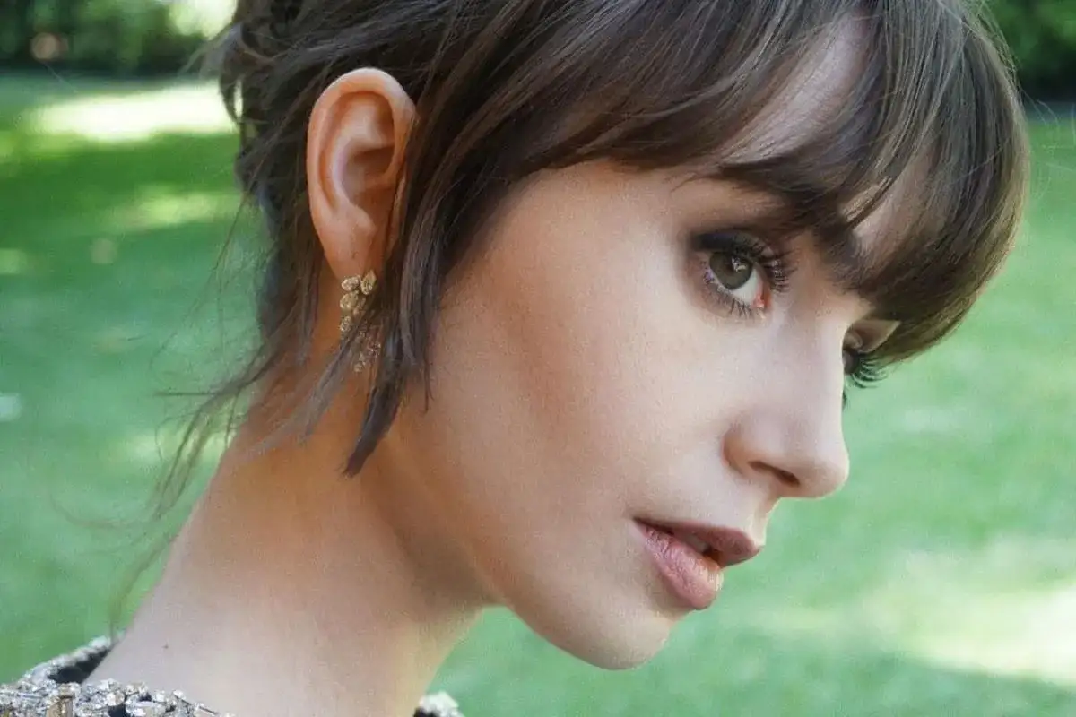Kobieta z **baby bangs** i delikatną biżuterią w uchu, na tle zieleni.