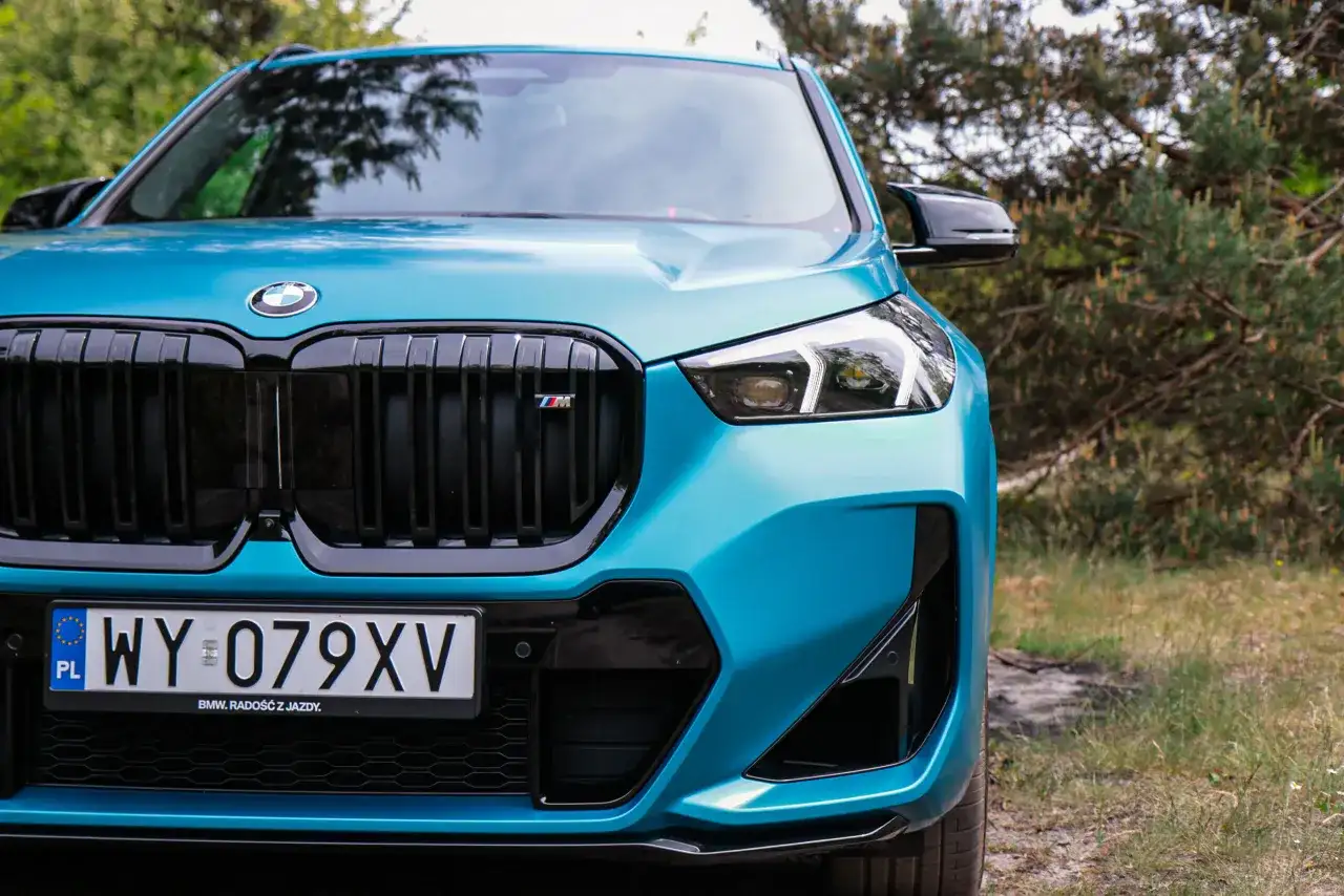 Turkusowy BMW X1 2024 z czarnym grillem i emblematem M, gotowy na przygodę w otoczeniu natury.