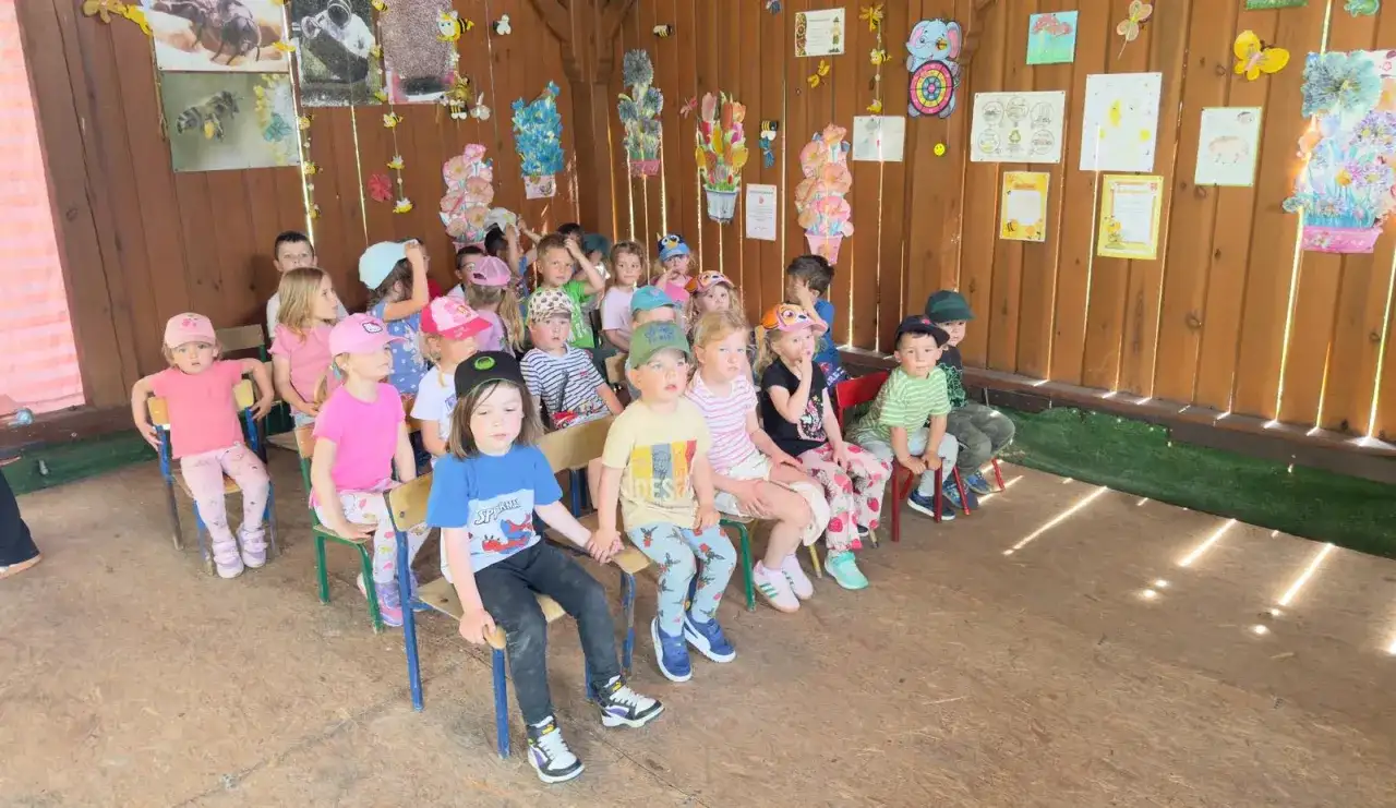 Dzieci siedzą na krzesłach w **zespole szkolno przedszkolnym**, słuchając uważnie.