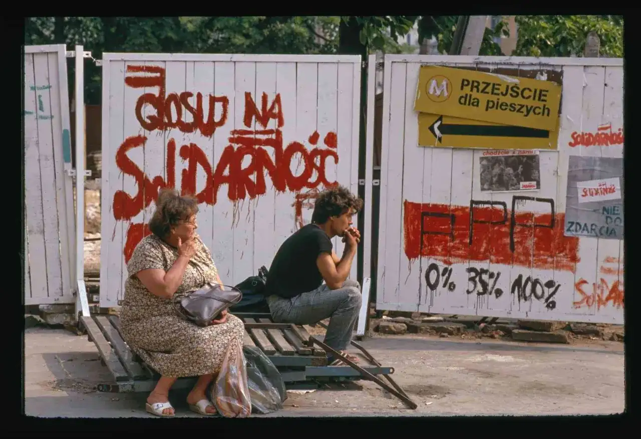 Dwie osoby siedzą na ławce przed płotem z napisem "Głosuj na Solidarność". To symboliczny obraz upadku komunizmu.