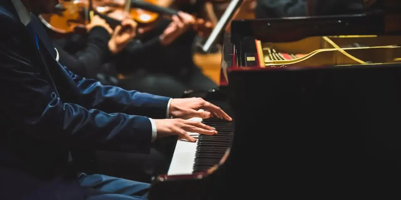 Pianista w eleganckim garniturze gra na fortepianie, a przed nim leżą nuty do nauki gry na pianinie. W tle orkiestra smyczkowa.