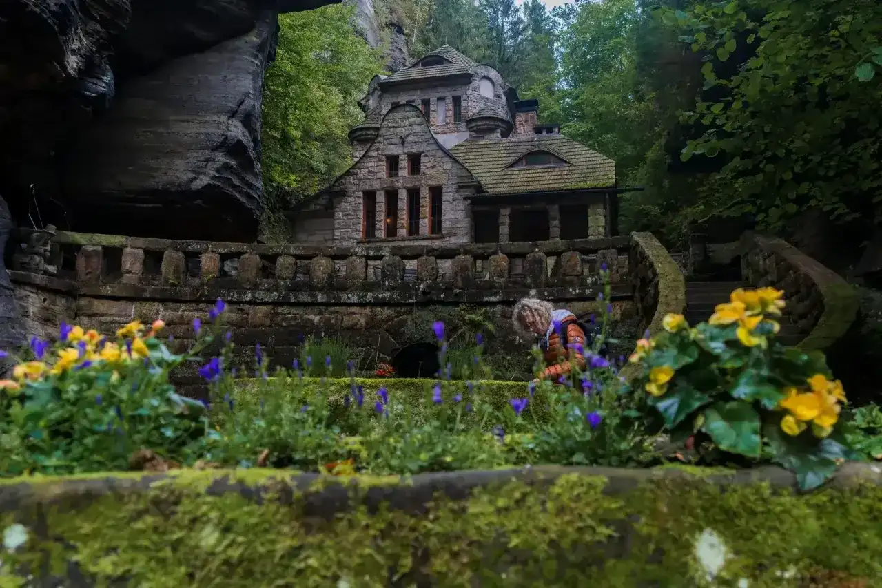 Kamienny domek w skalnym otoczeniu, idealne miejsce na czeska szwajcaria noclegi. Wokół kwitnące kwiaty i zielone drzewa.