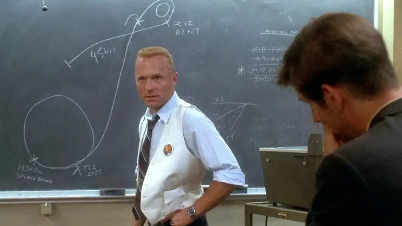 Gene Kranz, w białej kamizelce, omawia trajektorię misji Apollo 13 na tablicy.