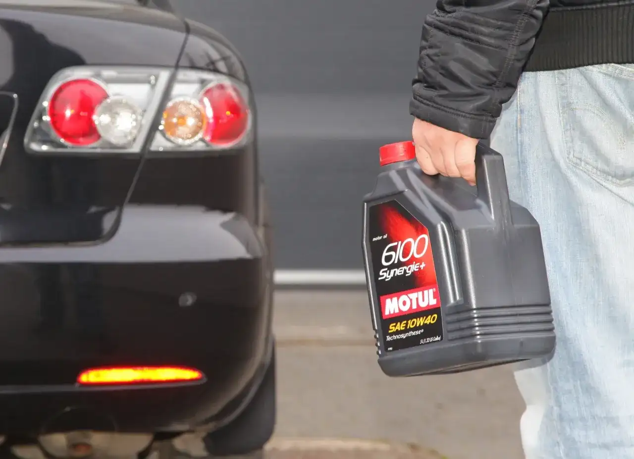 Osoba trzyma olej Motul 6100 Synergie+ SAE 10W40 obok tylnej części czarnego samochodu. Idealny wybór, by dobrze dobrać olej.