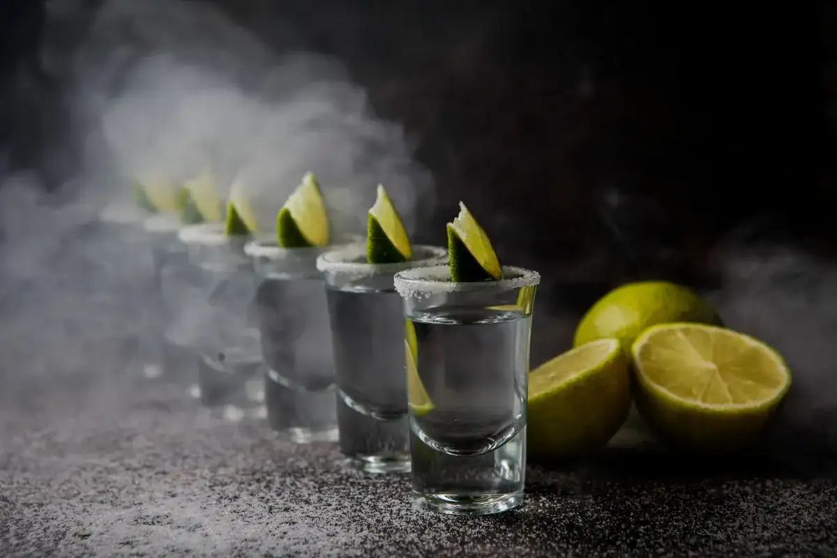 Rząd kieliszków z solą i limonką, wypełnionych przejrzystym płynem, otoczony mgłą. Obok leżą połówki limonki. Idealna sceneria na wieczór z tekilą.