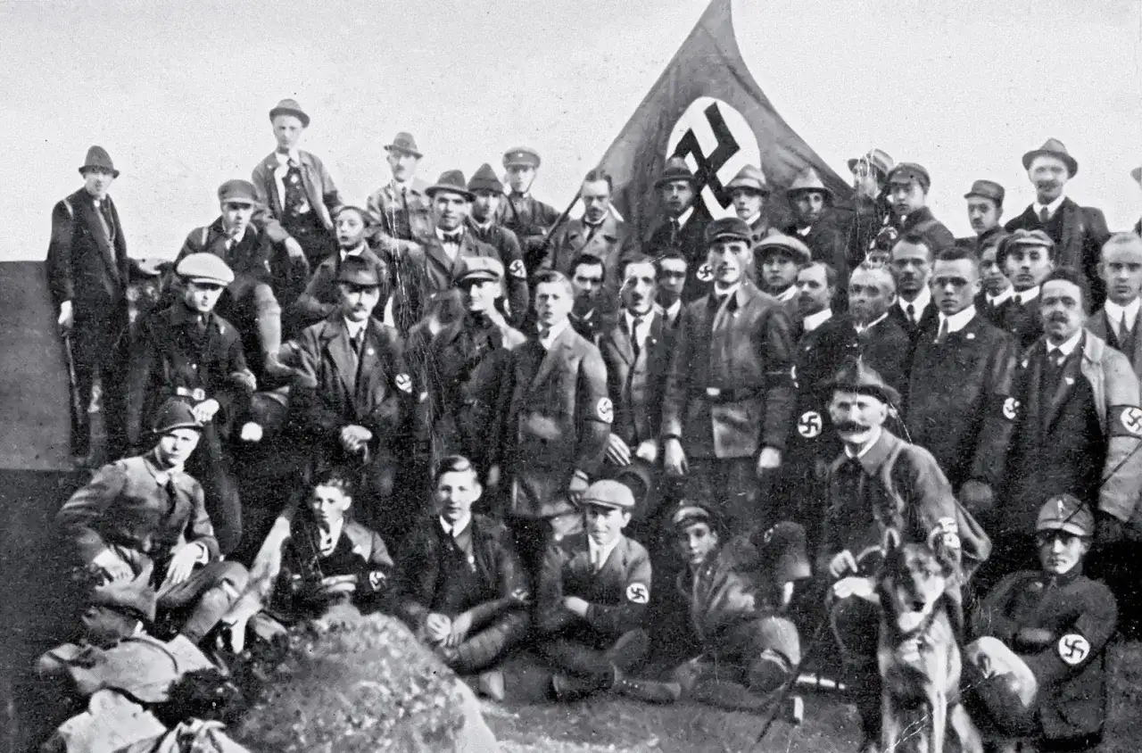 Grupa mężczyzn w mundurach i kapeluszach, z flagą NSDAP w tle. To zdjęcie pokazuje członków partii nazistowskiej, czyli NSDAP.