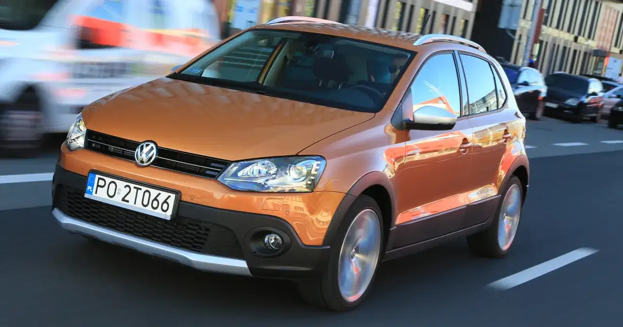 Pomarańczowy Volkswagen Polo Cross w ruchu miejskim, z widocznym kierowcą i innymi samochodami w tle.