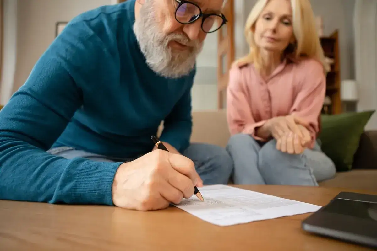 Starszy mężczyzna z brodą podpisuje dokumenty, podczas gdy kobieta siedzi obok. Rozmowa o dziedziczeniu.