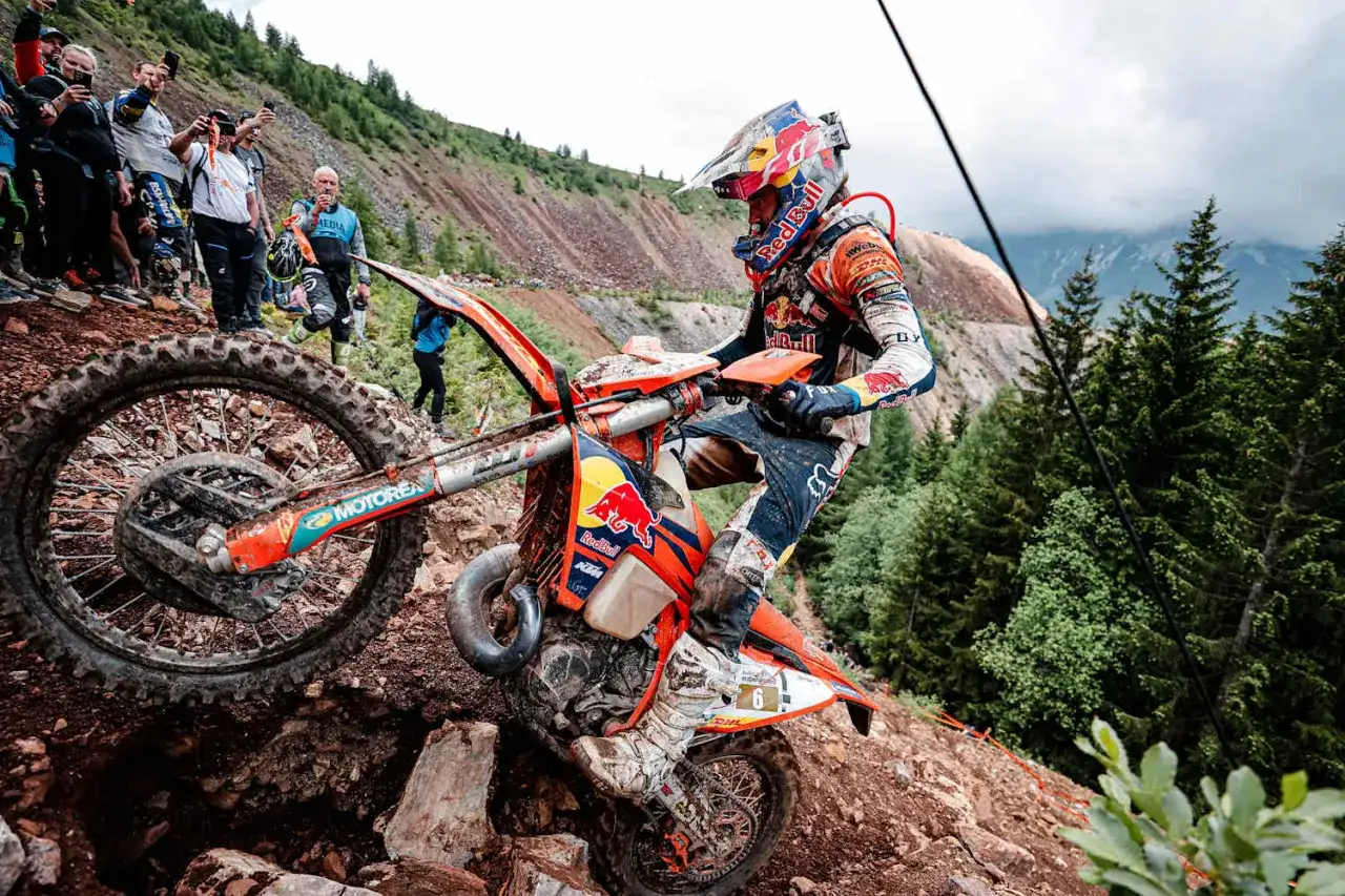 Motocyklista walczy z kamienistym podjazdem podczas Erzberg Rodeo. Widzowie obserwują zmagania na stromym zboczu.